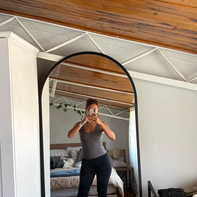 A woman in a gray tank top and dark leggings takes a mirror selfie with her phone in a bedroom with a wooden ceiling.