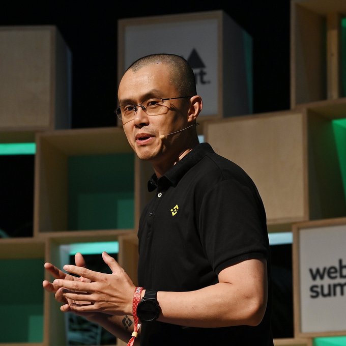 Changpeng Zhao (CZ), Binance's founder, speaks at the Web Summit while wearing a black polo shirt and glasses, with a visible "web summit" sign in the background. The image accompanies coverage of his remarks about allowing Hyperliquid to operate legally in the US as a gateway to broader decentralized finance services.