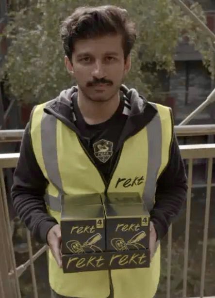 A man with a mustache wearing a yellow high-visibility vest labeled 'rekt' holds stacked black boxes marked 'rekt 4' in an outdoor setting.