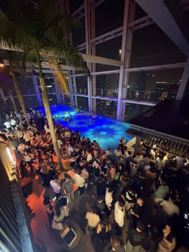 A large crowd of people gathers around a rooftop swimming pool at night in a high-rise building.