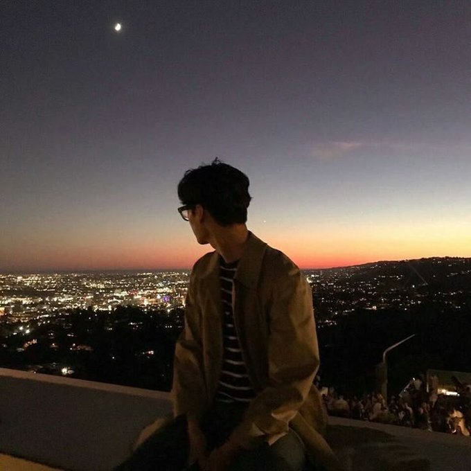 iced tea with a straw, dressed casually, sits on a rooftop at sunset, overlooking the city skyline and a crescent moon.