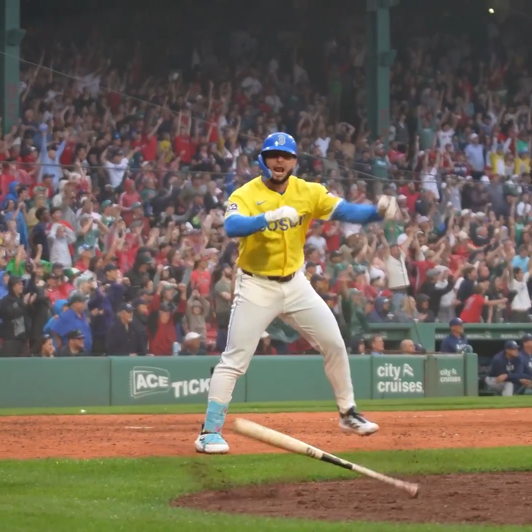 A Boston Red Sox baseball player in a yellow jersey celebrating at home plate with a bat dropped on the ground, while a cheering crowd raises their arms in the stands at Fenway Park.