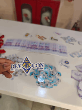 A hand holds a Devcon India sticker above a table covered in Devcon-branded materials and artwork. The text "DevCon India" is prominently displayed on the sticker, aligning with the post about the community gathering ahead of Devcon VIII in India.