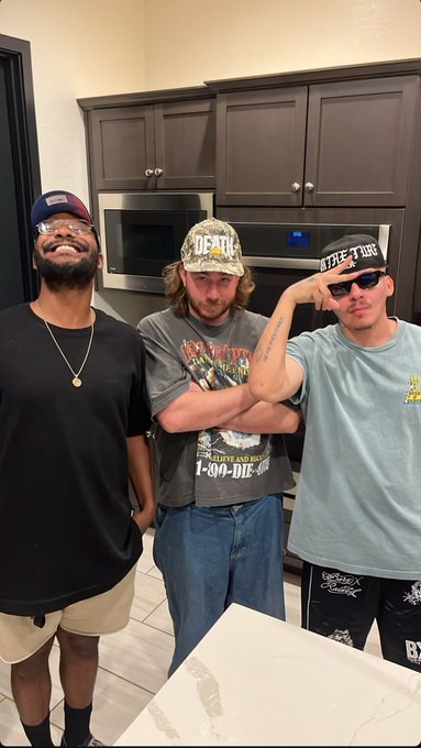 Three men pose in a kitchen; one wears a 'DEATH' cap, another wears a 'LIVE FAST DIE' shirt, and the third wears a cap with sunglasses.