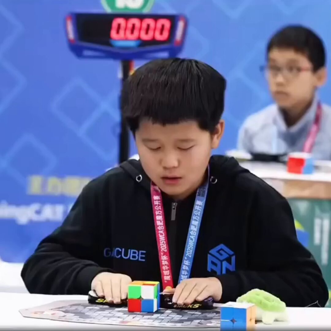 The image shows a young boy sitting at a table, playing with Rubik's cubes. He is wearing a black hoodie that has "rubik cube" written on it. The boy appears to be focused on solving the puzzle before him. In front of him are several Rubik's cubes in various colors and positions, indicating he is actively engaged in the activity.
