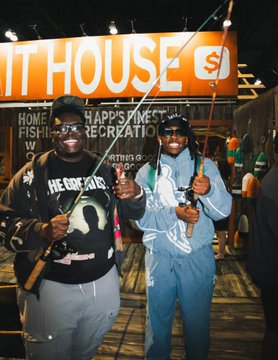 Two men in costumes hold fishing rods in front of an orange "BIT HOUSE" booth with text reading "HOME OF FISHING'S HAPPIEST RECREATION," fitting the post about arriving for fishing and leaving with bitcoin.