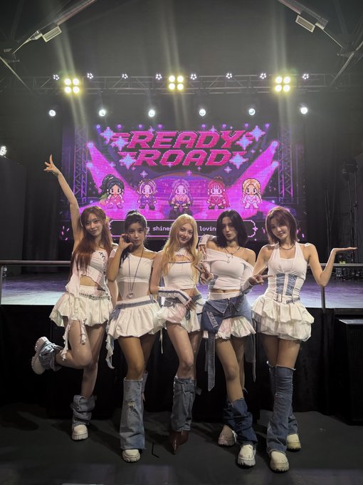 A K-pop girl group photo of five female performers in coordinated white tank tops, ruffled skirts, and denim boots posing on stage in front of a 'READY, ROAD' LED screen displaying anime-style character avatars.