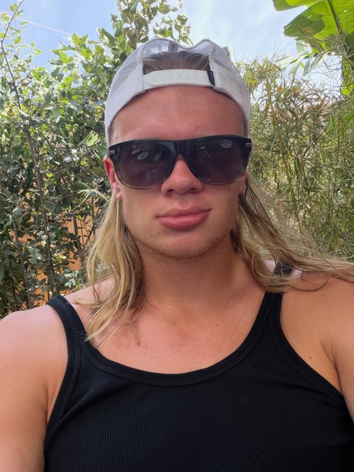 A selfie of a man with long blonde hair wearing a white backwards cap, sunglasses, and a black tank top, with green foliage behind him.