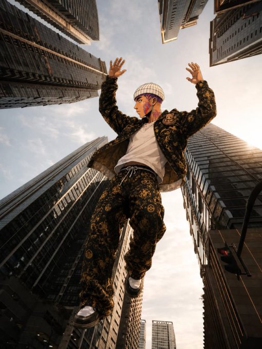 A low-angle shot of a person wearing a black and gold patterned outfit and mask, posing with arms raised between tall skyscrapers.