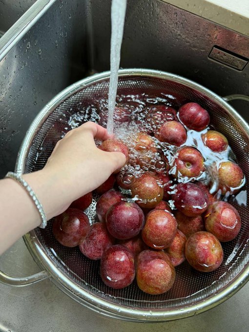 The image shows a person washing red plums in a sink, with water coming out of the faucet. The scene appears to be set in an office environment, as there are several other people visible in the background who seem to be working or engaged in their tasks.
