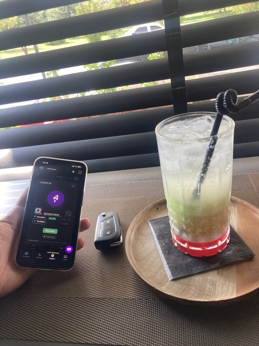 A hand holds a smartphone displaying a "BOSSIPGIRL WOW" profile on a social media app, next to a car key fob and a glass of a green, ice-filled beverage with a black curly straw on a wooden tray.