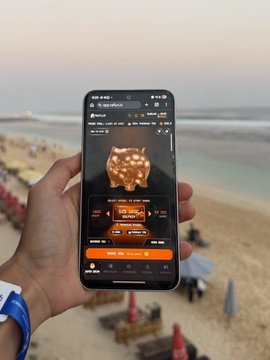 A hand holding a smartphone displaying a cryptocurrency trading app with an orange/pig-themed NFT, set against a beach backdrop.