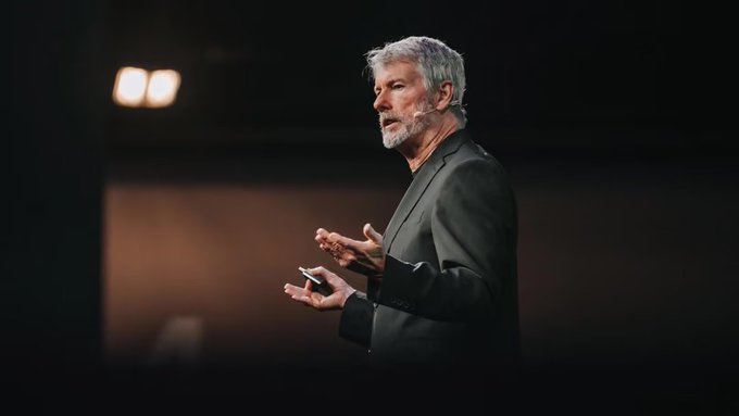 A man with grey hair and beard, wearing a dark blazer, gestures while speaking on a dimly lit stage, possibly giving a presentation.