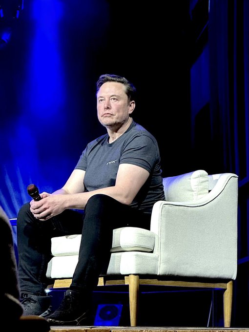 Elon Musk sits on a stage in a white armchair, wearing a dark "X" branded t-shirt and holding a microphone, appearing contemplative as his net worth of $879.2 billion brings him within reach of trillionaire status.