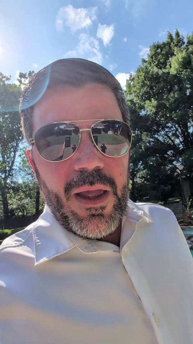 The image features a man wearing sunglasses, a white shirt, and a beard. He appears to be posing for a photo or video update with a caption that reads "Atentos a $MSTR y $BTC." The background of the image is blurred, but there are trees visible in the distance.