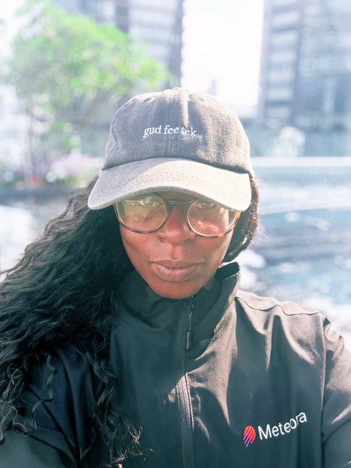 A person wearing a 'gud fee tek' cap, sunglasses, and a Meteora jacket standing outdoors.