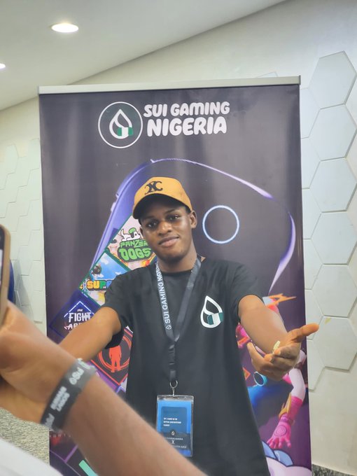 A young man wearing a black 'SUI GAMING NIGERIA' t-shirt and a tan cap posing in front of a promotional banner with the same branding.