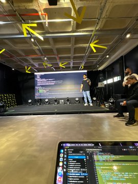 A presentation stage setup with yellow arrows pointing to overhead equipment, a presenter on stage, and a laptop in the foreground showing a video editing or streaming interface.