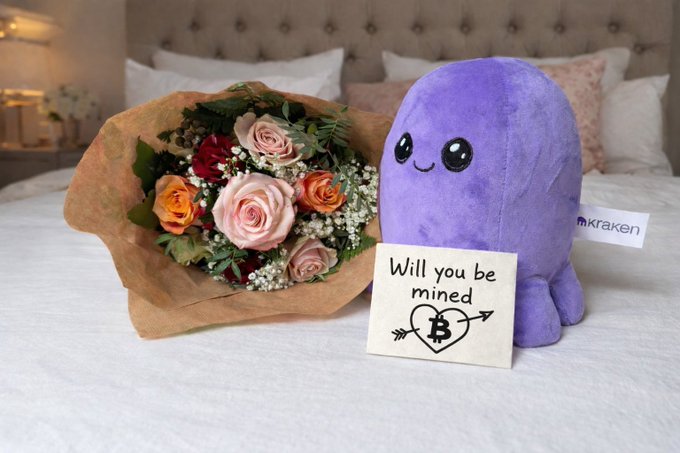 A Kraken-branded Valentine's gift bundle on a bed, featuring a bouquet of roses, a purple octopus plush, and a card reading 'Will you be mined' with a Bitcoin heart and arrow design.