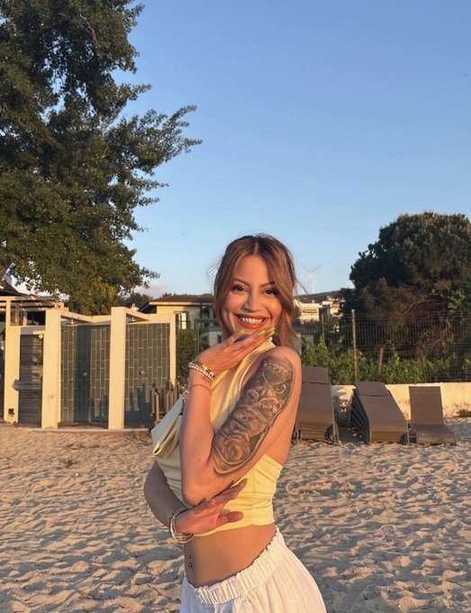A smiling young woman with a detailed rose tattoo sleeve poses on a sandy beach at golden hour, wearing a yellow top and white skirt, with beach cabanas and lounge chairs in the background. The "gm" (good morning) caption matches the bright, cheerful morning vibes of her sunny beachside photo.