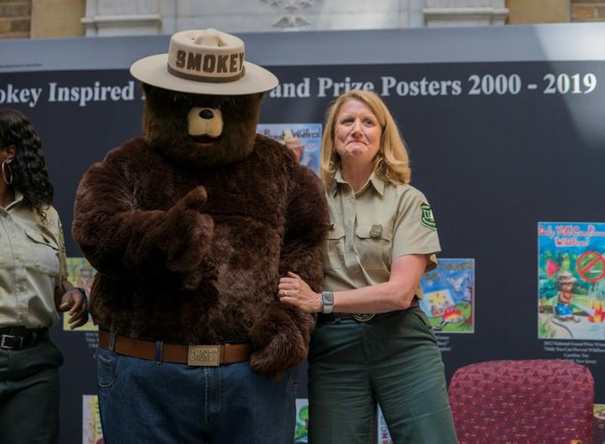 Smokey Bear mascot wearing a hat and belt, standing next to a woman in a National Park Service uniform in front of an exhibit about 'Smokey Inspired' posters.
