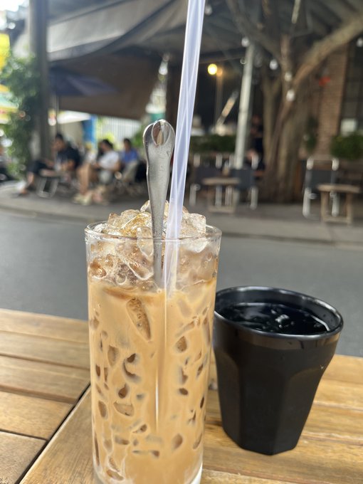 A glass of iced milk coffee and a small black ceramic cup on a wooden outdoor café table, with blurred patrons and trees visible in the background.
