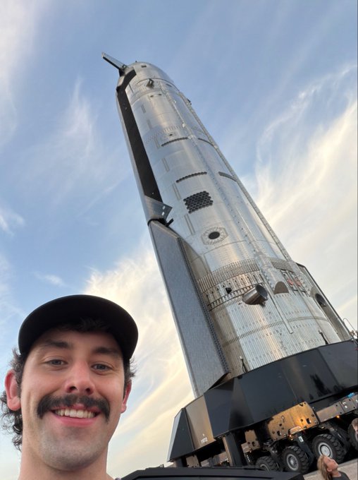 A man with a mustache and cap takes a selfie in front of SpaceX's towering Starship rocket against a partly cloudy sky, beaming with excitement as the massive spacecraft rises behind him. The image captures his joy and enthusiasm for SpaceX's next-generation launch vehicle, embodying the post's sentiment that "Starship makes me happy."