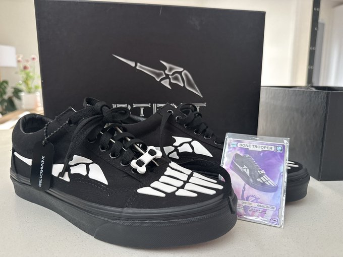A pair of black skate shoes with white skeletal hand and wing graphics, displayed in front of a black box with a silver wing logo, accompanied by a trading card labeled 'BONE TROOPER'.