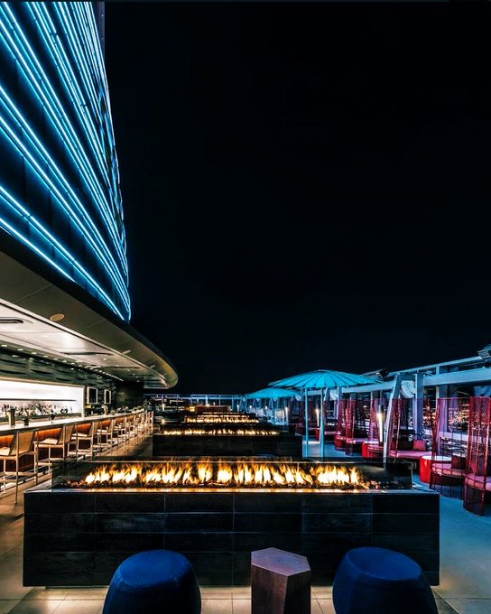 A modern rooftop lounge at night, featuring a long linear fireplace, blue neon light strips on the building facade, and seating overlooking a city.