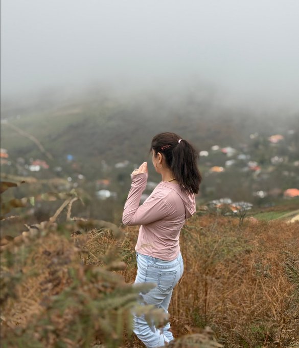 A woman in a pink hoodie and jeans stands in a grassy hillside looking out at a misty town in the valley below.