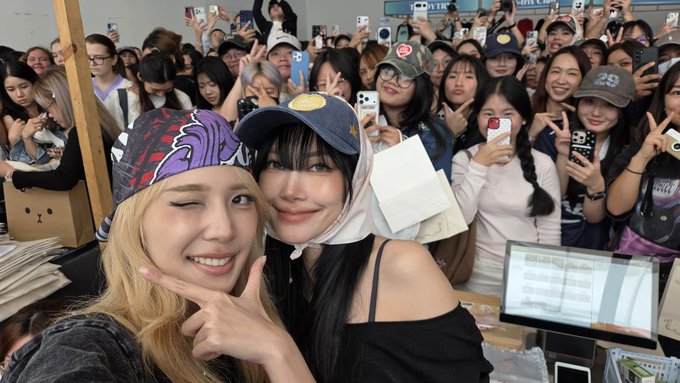 Two women pose for a selfie at what appears to be a fan meet-and-greet, surrounded by a crowd of enthusiastic fans holding up phones to take photos; one wears a bandana with purple lettering while the other sports a black cap with a gold emblem. The scene aligns with the post about meeting fans (แฟนคลับ/กะลายน้ำ) for the "Mint Awards 2026 x JJJ" event, as the excited crowd and selfie-style photo capture the celebratory fan interaction moment.