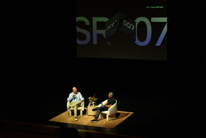 Two men sit on a small stage in a dimly lit auditorium during the a16z Speedrun SR007 founders event, with a large projection screen displaying "SR007" and "007" behind them. The setting appears to be a fireside chat or panel discussion, aligning with the post's mention of gathering founders of the Speedrun accelerator batch.