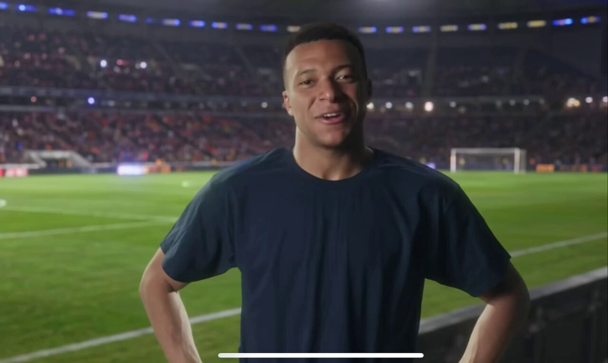 The image features a man wearing a blue shirt, standing in front of an empty stadium with a soccer field behind him. The man appears to be smiling for the camera, possibly posing as a soccer player or a sports enthusiast. The background shows that there are no other people visible at this moment.