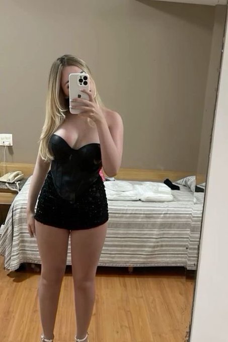 A blonde woman in a black corset and sequined mini skirt takes a mirror selfie in a bedroom, holding up an iPhone to obscure her face. The room features a bed with striped bedding and a wooden floor in the background.