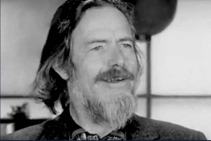 A black-and-white close-up of a bearded man speaking indoors, with a microphone visible near the upper right.