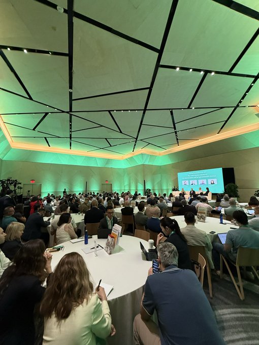 A packed conference room at the RWA Summit in NYC glows with green ambient lighting as attendees seated at round tables watch a presentation on "The Coming Revolution: Real World Assets & Onchain Finance" displayed on a large screen. The scene captures the strong turnout and engaged audience for the onchain finance event mentioned in the post.