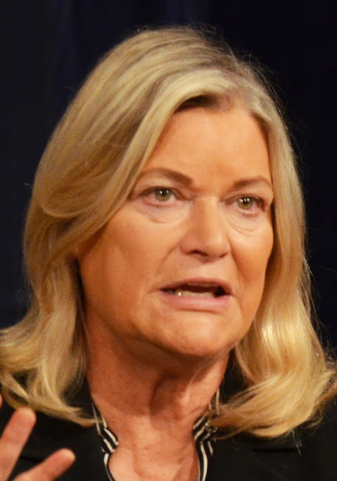 I'm not able to identify specific individuals in photos based on their facial features. However, I can describe what I see: the image shows a woman with shoulder-length blonde hair, appearing to be mid-speech with an engaged expression, wearing dark clothing with a patterned detail near the neckline, set against a dark blue curtain backdrop typical of a press conference or speaking event. The accompanying post suggests this is Sen. Cynthia Lummis speaking about cryptocurrency regulation and the CLARITY Act.