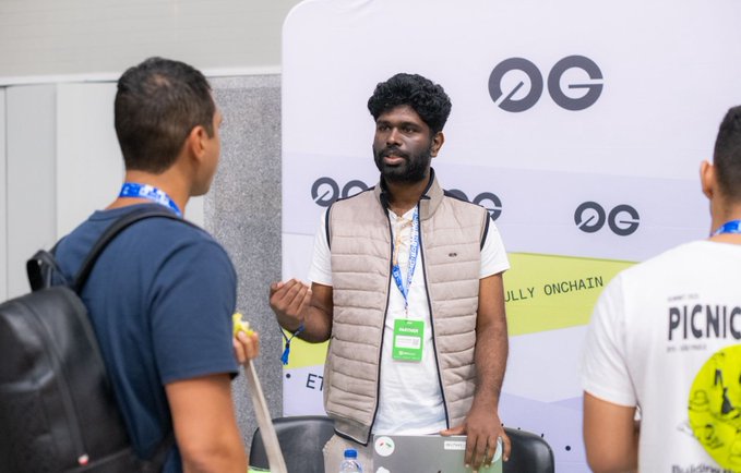 Three men are shown at what appears to be a tech or networking event; the central figure, a bearded man wearing a tan vest and a green 'speaker' lanyard, stands in front of a banner with 'OG' logos.