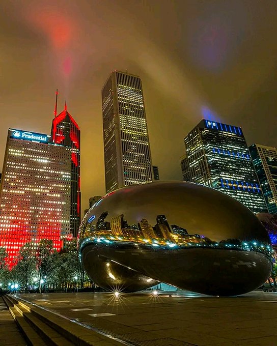 The image shows an urban city skyline at night with skyscrapers, including some lit up with red lights, surrounded by trees that add greenery to the scene. The focal point of the photograph is a large silver sculpture or object resembling a shell. This piece of art appears to be located in front of a plaza and is situated within a public square. A post on this city landmark captures attention due to its unique design and its representation of cross-chain infrastructure, highlighting the importance of liquidity and efficient asset management across ecosystems.