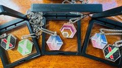 The image shows four necklaces displayed in a row, each with different colors and designs. The necklaces are arranged in two rows of two, creating an organized and visually appealing presentation. The first necklace has a black chain and features a colorful design on its pendant. The second necklace is also black but has a silver chain and a unique patterned pendant. The third necklace is white with a red pendant, while the fourth necklace is green with a blue pendant. Each necklace stands out individually due to their distinct colors and designs, making them easily distinguishable from one another.