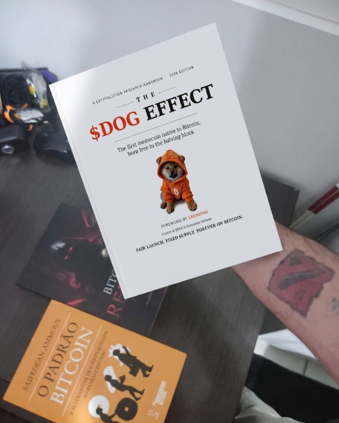 A hand holds up a paperback book titled 'The $DOG Effect' (with 'A Cryptocurrency Research Handbook — 2026 Edition' and subtitle 'The first memecoin native to Bitcoin, born free in the halving block'), alongside another book titled 'O Padrão Bitcoin' by Saifedean Ammous visible on the desk below.