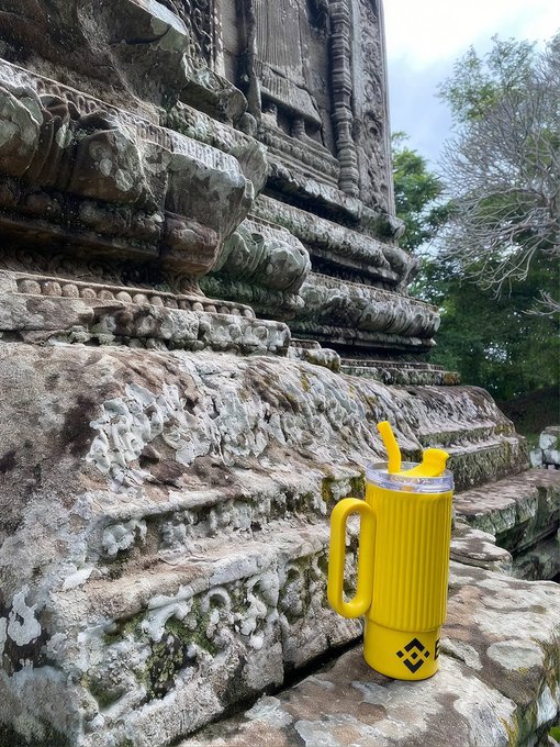 A modern Binance-branded yellow tumbler sits incongruously on the weathered stone steps of an ancient temple ruin, contrasting the cryptocrurrency craze with centuries-old history. The tumbler displays the recognizable Binance logo and diamond symbol.
