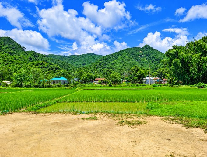 A scenic landscape shows lush green rice paddies and small village houses nestled at the foot of forested mountains under a bright blue, cloudy sky.