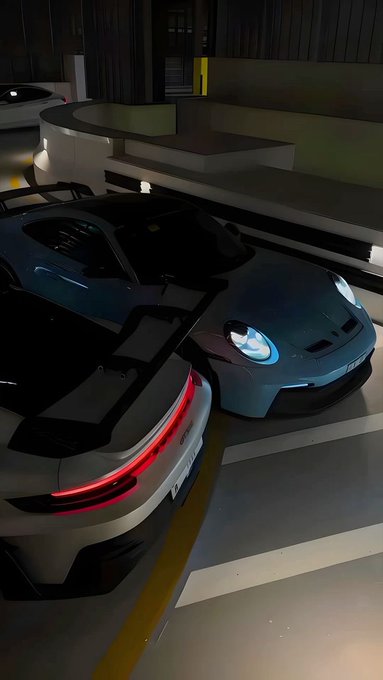 Two high-performance sports cars, one white with glowing red taillights and one dark grey with illuminated blue headlights, are parked side by side in a dimly lit underground garage.