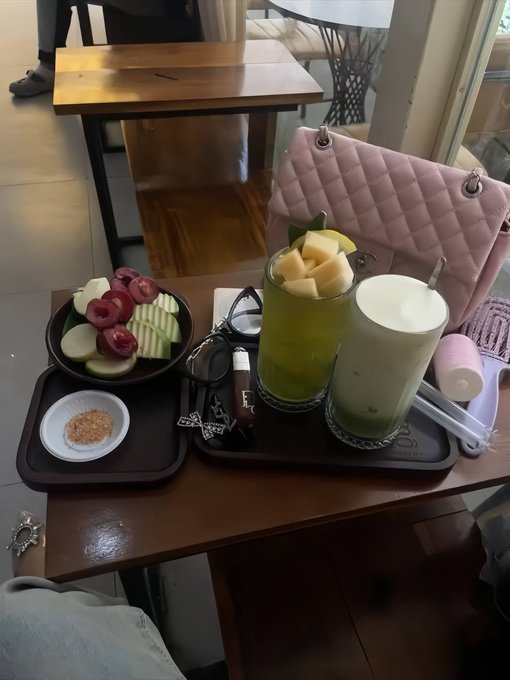 The image shows a dining table with various items on it, including two glasses of drinks, a bowl filled with fruit (apples), and a purse sitting next to them. The scene appears to be set up for a meal or gathering, possibly in a cafe or restaurant setting.