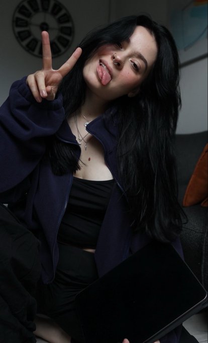 A young woman with long dark hair makes a peace sign and sticks out her tongue while wearing a purple jacket and black top, sitting indoors with a clock on the wall behind her.