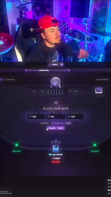 The image shows a man wearing a red baseball cap sitting behind a computer screen, playing a video game with a purple background. The man appears to be focused on his gameplay while wearing a hat that matches the color of the background. In the bottom right corner of the image, there is a post about a new game called "Interact & Drop Sol wallets". This post seems to be related to the gaming session taking place in front of the man with the red cap.