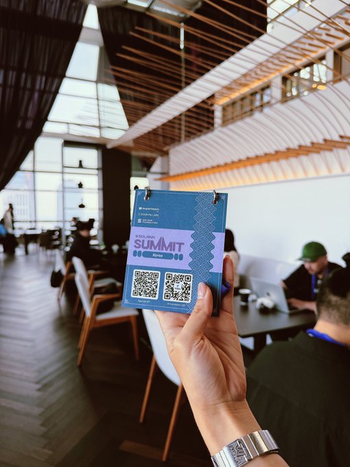A hand holds up a blue "Decrypt Summit Korea" badge with two QR codes at what appears to be a modern venue, while attendees work on laptops at tables in the background—capturing the GM greeting from the Seoul-hosted crypto summit.