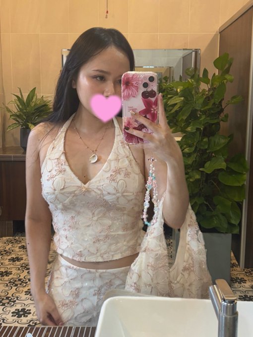 A mirror selfie of a woman in a white floral outfit holding a pink floral phone case, with a pink heart emoji covering her face.