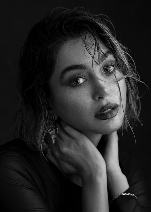 A black and white portrait photo of a young woman with short wavy hair, freckles, and bold lipstick, resting her chin on her hands.
